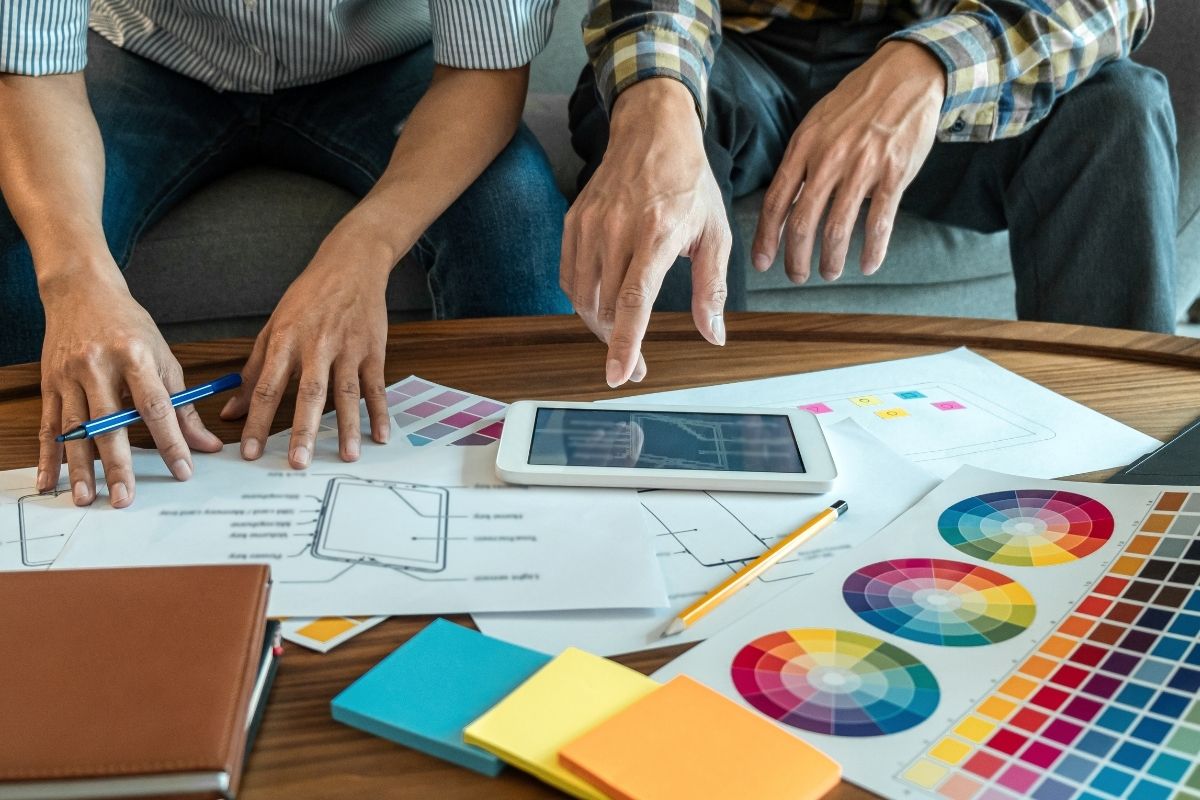 Hands of two designers reviewing wireframes and colour palettes around a tablet on a coffee table